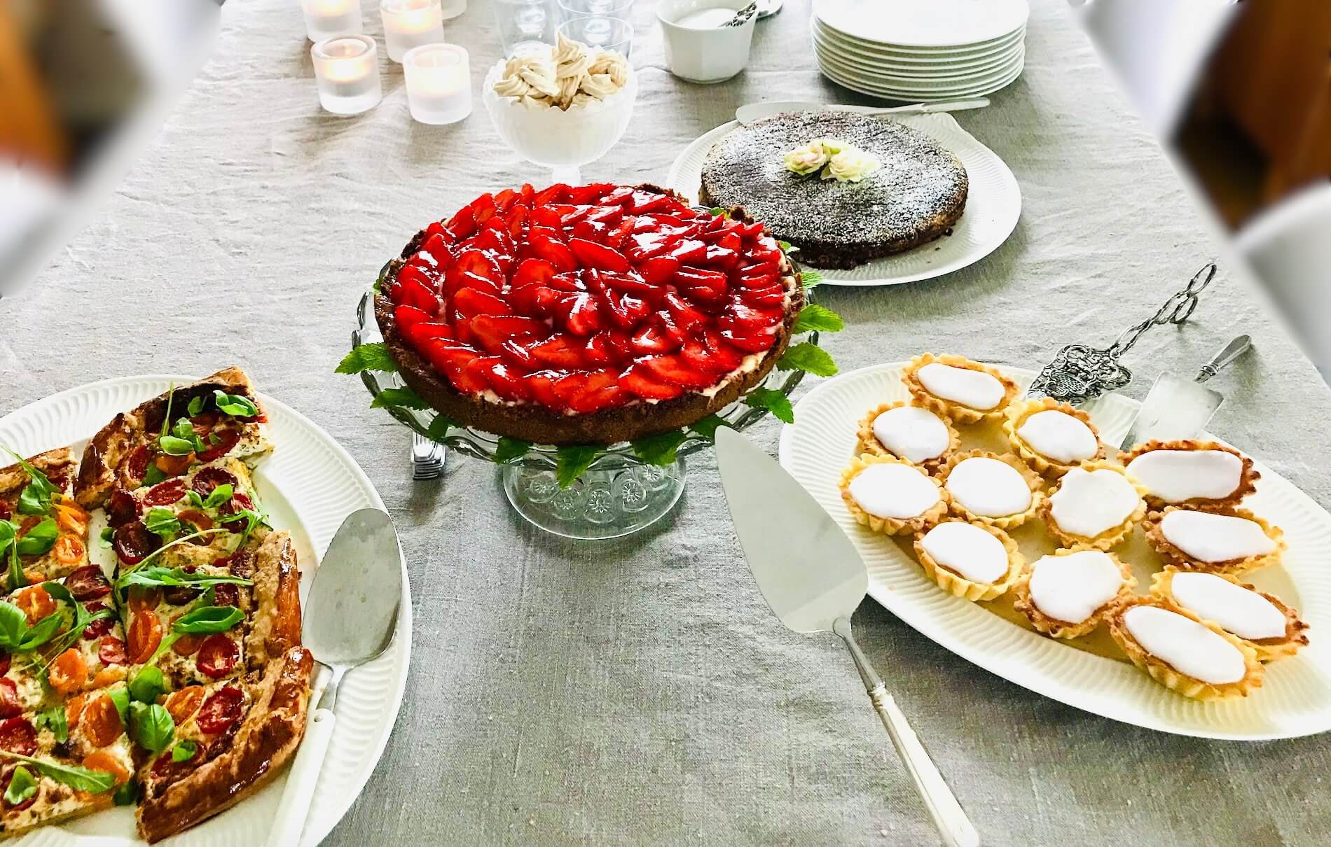 Finnish spring buffet dessert table featuring strawberry tart, mazarin pastries, chocolate mud cake, meringue cookies, and savoury tomato tart on a white linen table setting.