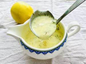 Creamy lemon dill sauce with chopped fresh dill, dripping from a sauce spoon into a porcelain saucer.