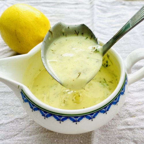 Creamy lemon dill sauce with chopped fresh dill, dripping from a sauce spoon into a porcelain saucer.