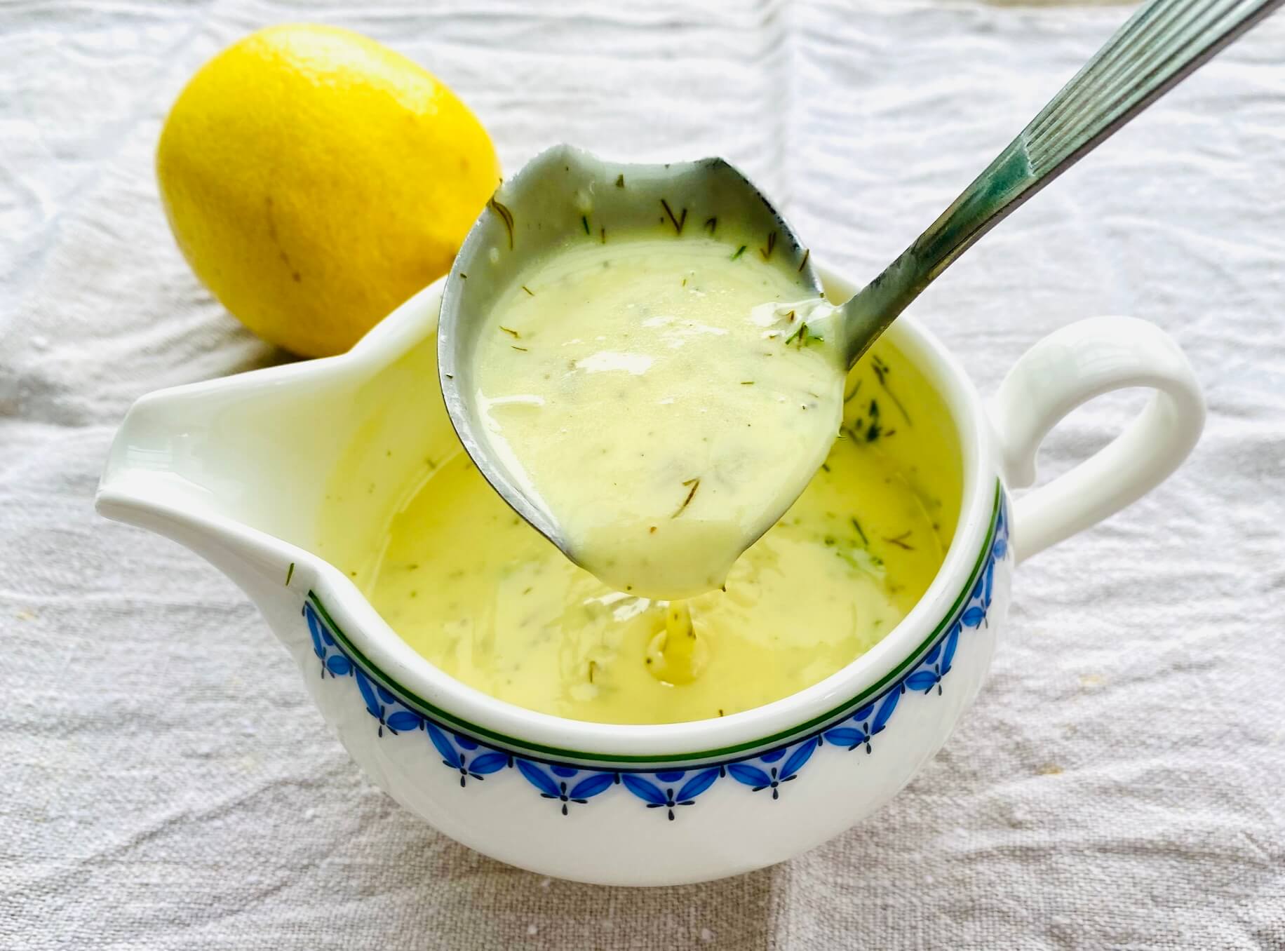 Creamy lemon dill sauce with chopped fresh dill, dripping from a sauce spoon into a porcelain saucer.