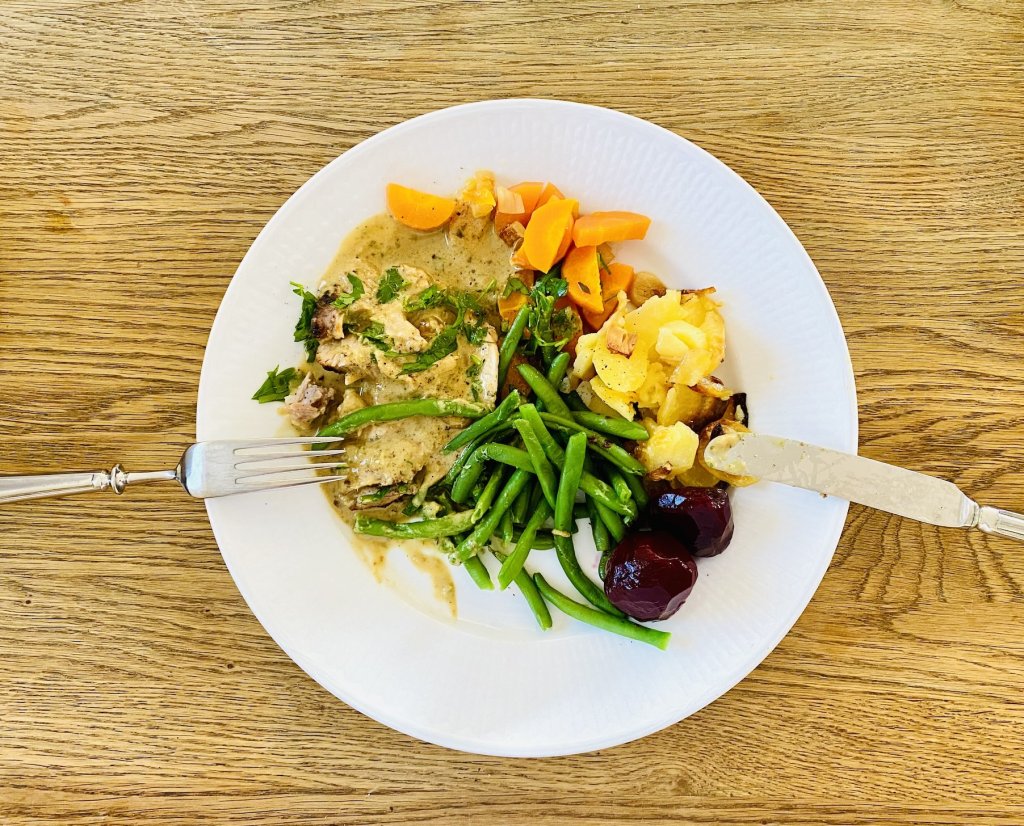 Sliced oven-baked pork with cream sauce, garlic potatoes, green beans, carrots, and pickled beetroot on a plate.