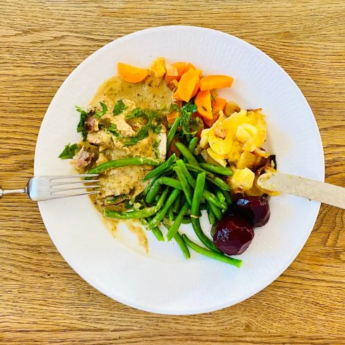 Sliced roast pork with creamy parsley sauce, garlic potatoes, green beans, carrots, and pickled beetroot on a plate.