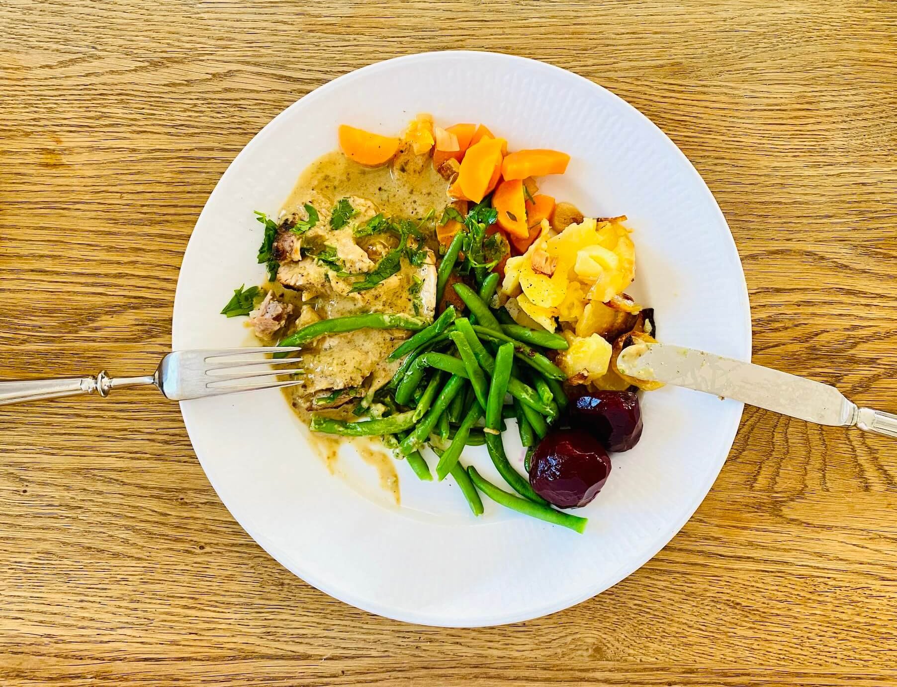Sliced roast pork with creamy parsley sauce, garlic potatoes, green beans, carrots, and pickled beetroot on a plate.